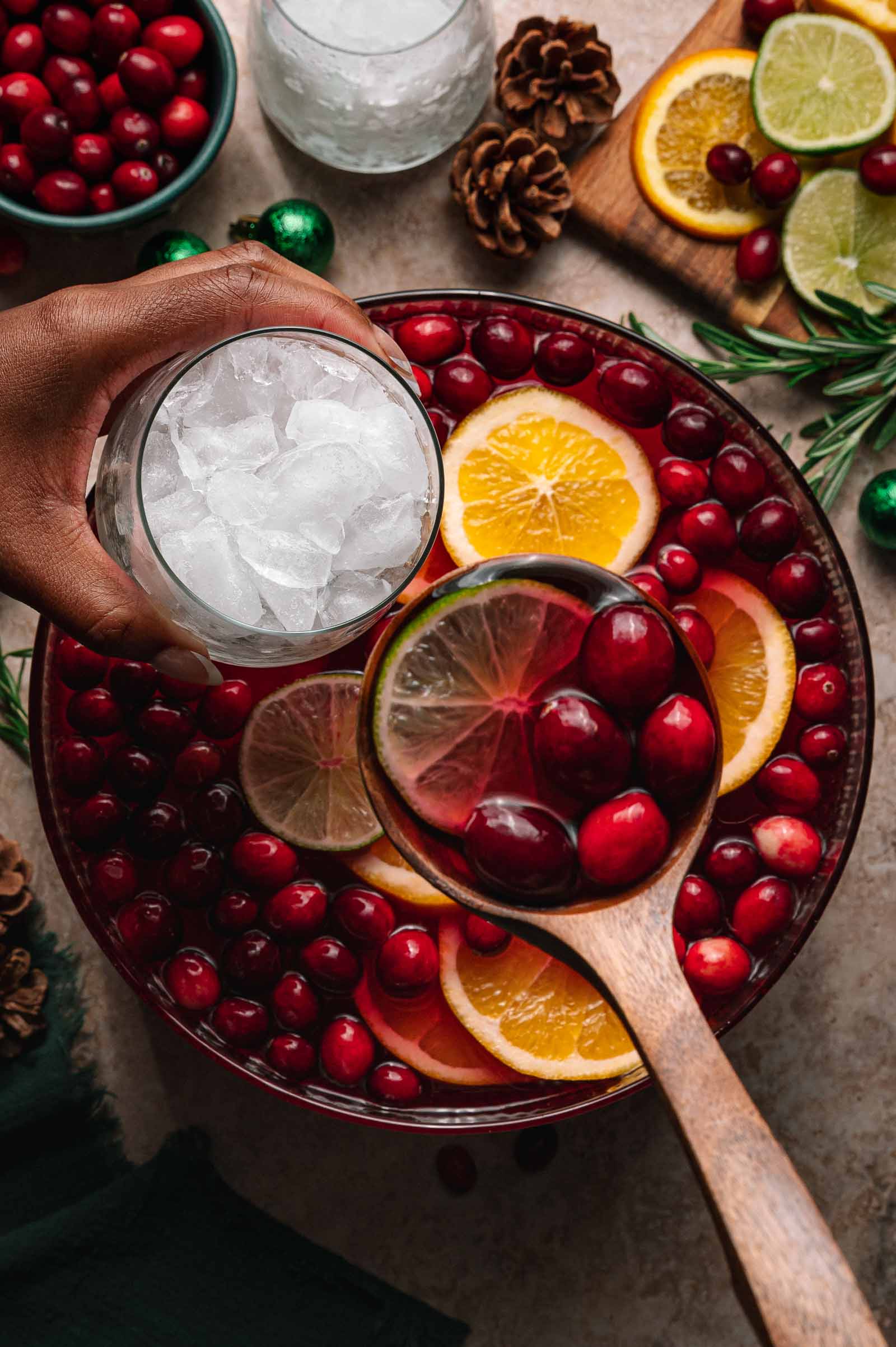 A hand holds a glass of ice over a bowl of jingle juice punch with cranberries, orange and lime slices as a ladle scoops some out.