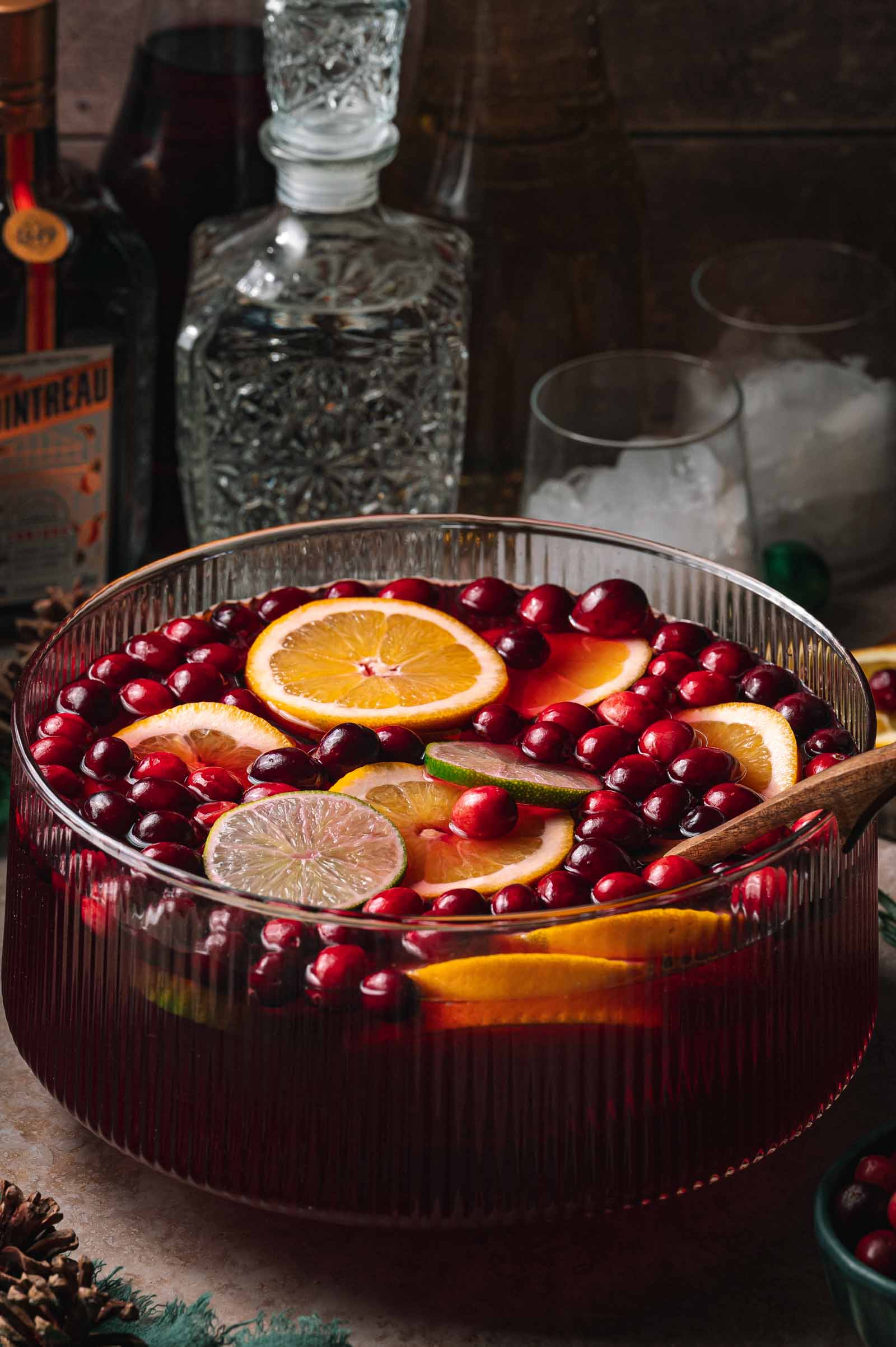 A glass punch bowl with red jingle juice batch cocktail, orange and lime slices, cranberries, and a wooden ladle; bottles and glasses behind.