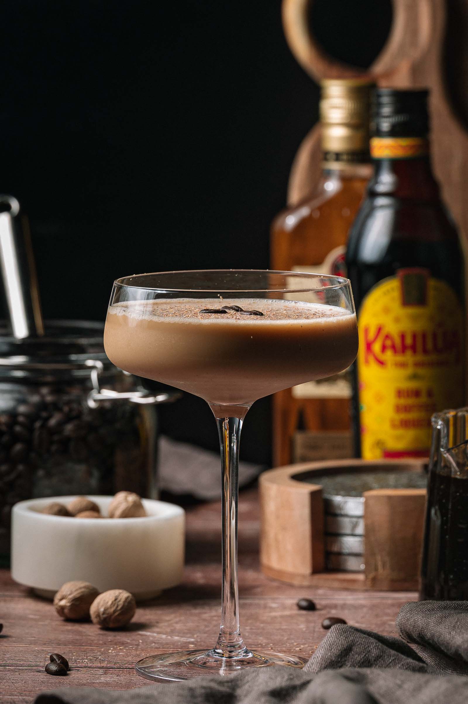 An eggnog espresso martini on a table is surrounded by coffee beans, nutmeg, and bottles of Kahlua and liquor in the background.