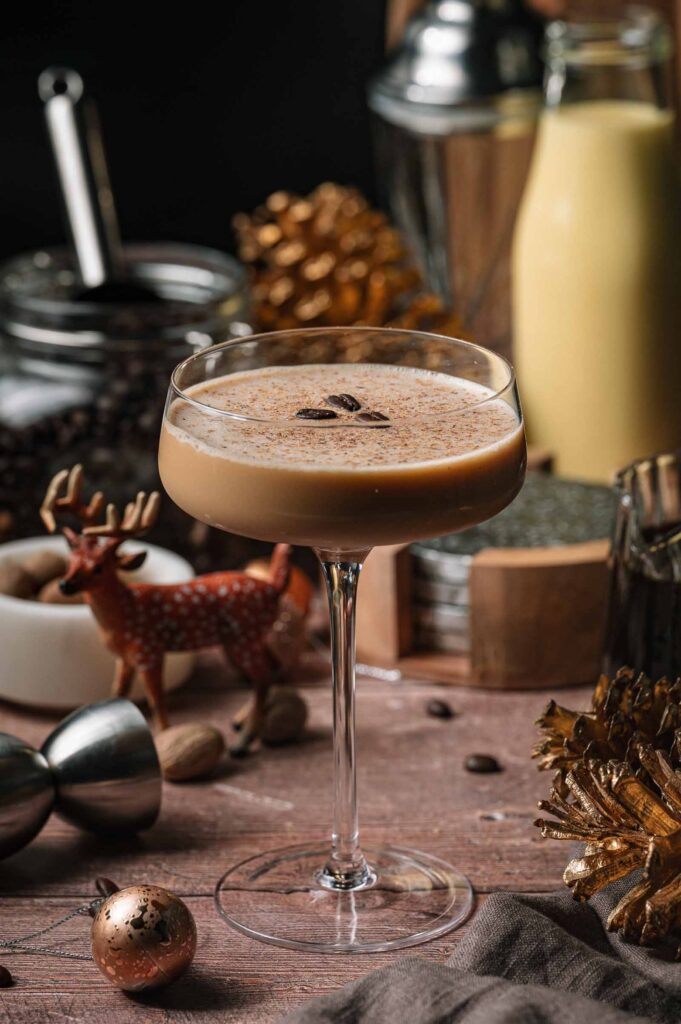 A creamy eggnog martni with coffee beans in a coupe glass rests on a wooden table amid festive d&eacute;cor and drink ingredients.