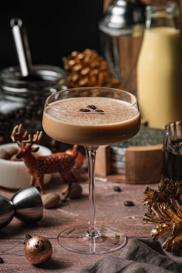 A creamy eggnog martni with coffee beans in a coupe glass rests on a wooden table amid festive décor and drink ingredients.