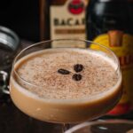 Eggnog espresso martini in a coupe glass with three coffee beans, liquor bottles blurred in the background.