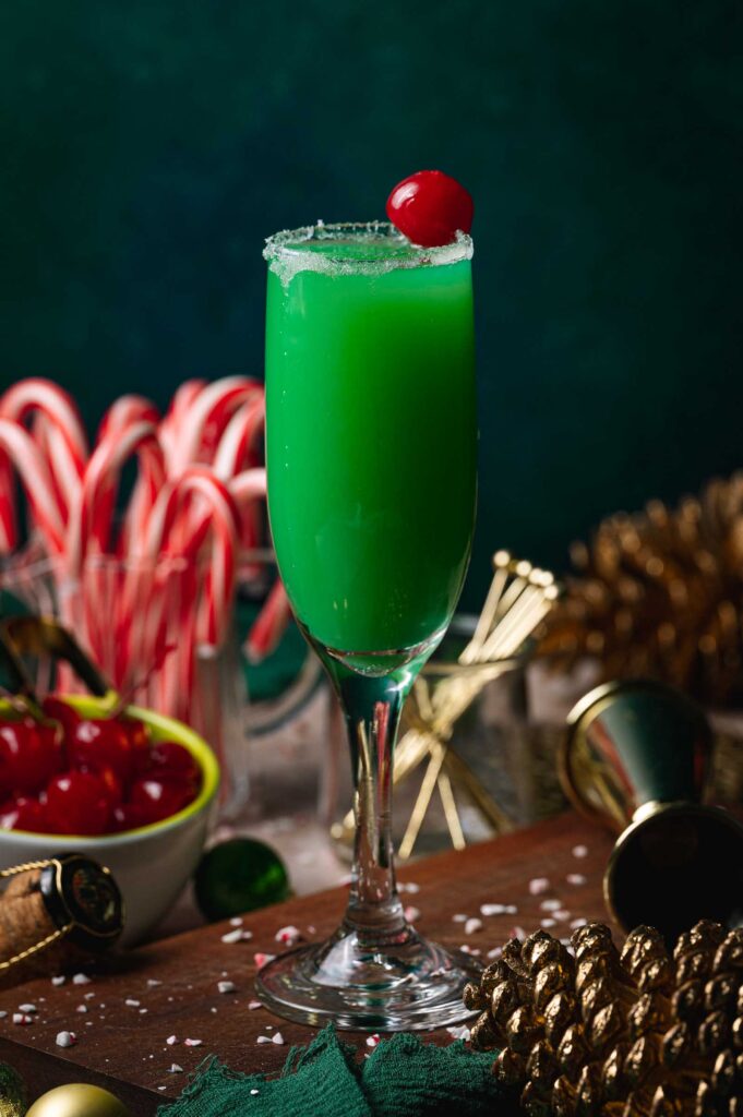 A tall festive Grinch mimosa cocktail with sugar rim and cherry sits on a festive table with candy canes, cherries, pinecones, and decorations.
