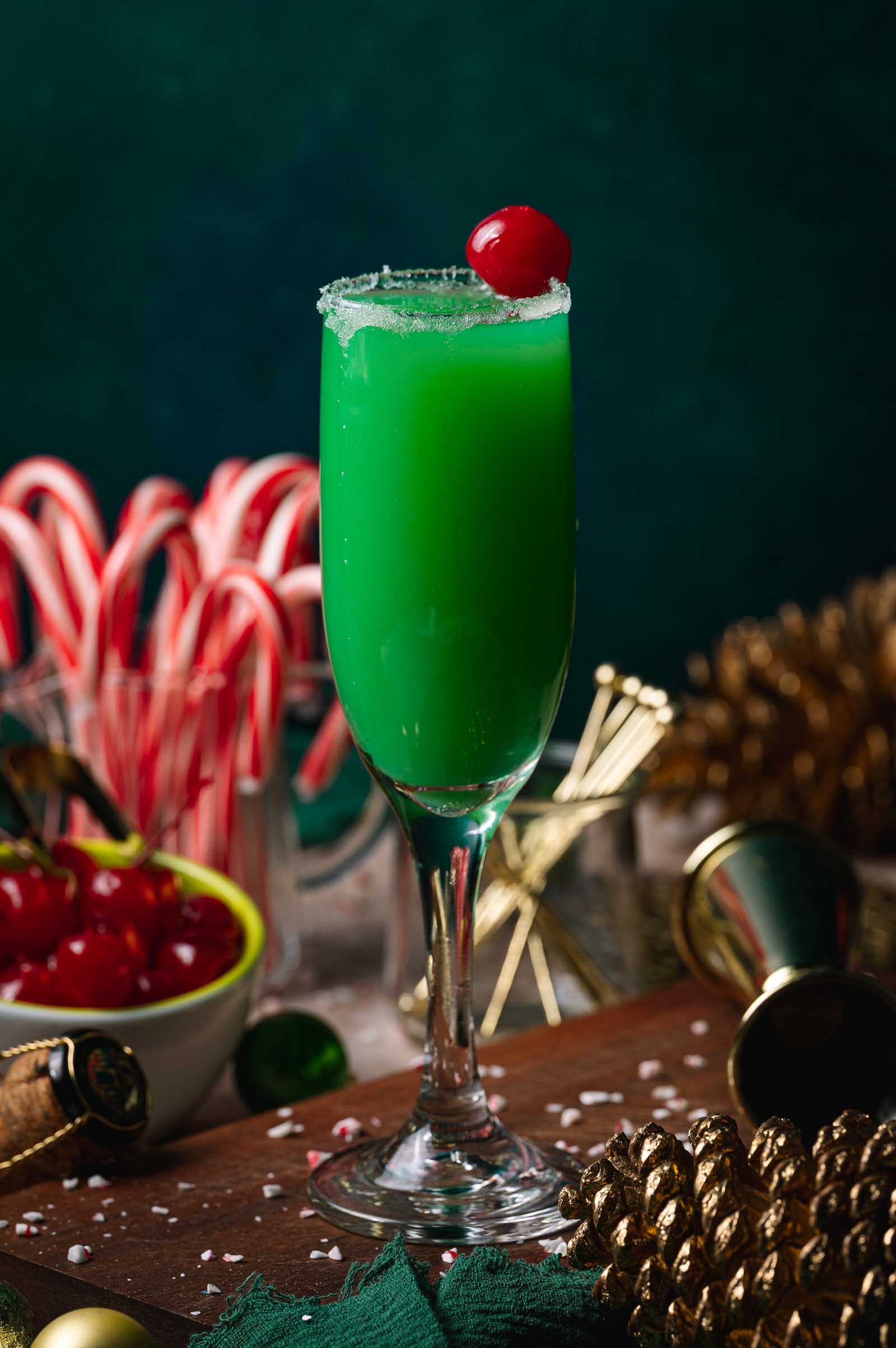 A tall festive Grinch mimosa cocktail with sugar rim and cherry sits on a festive table with candy canes, cherries, pinecones, and decorations.