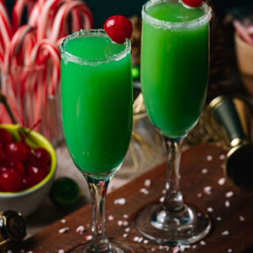Two Grinch mimosa cocktails with sugar rims and cherries sit in champagne flutes on wood, surrounded by candy canes and festive decor.