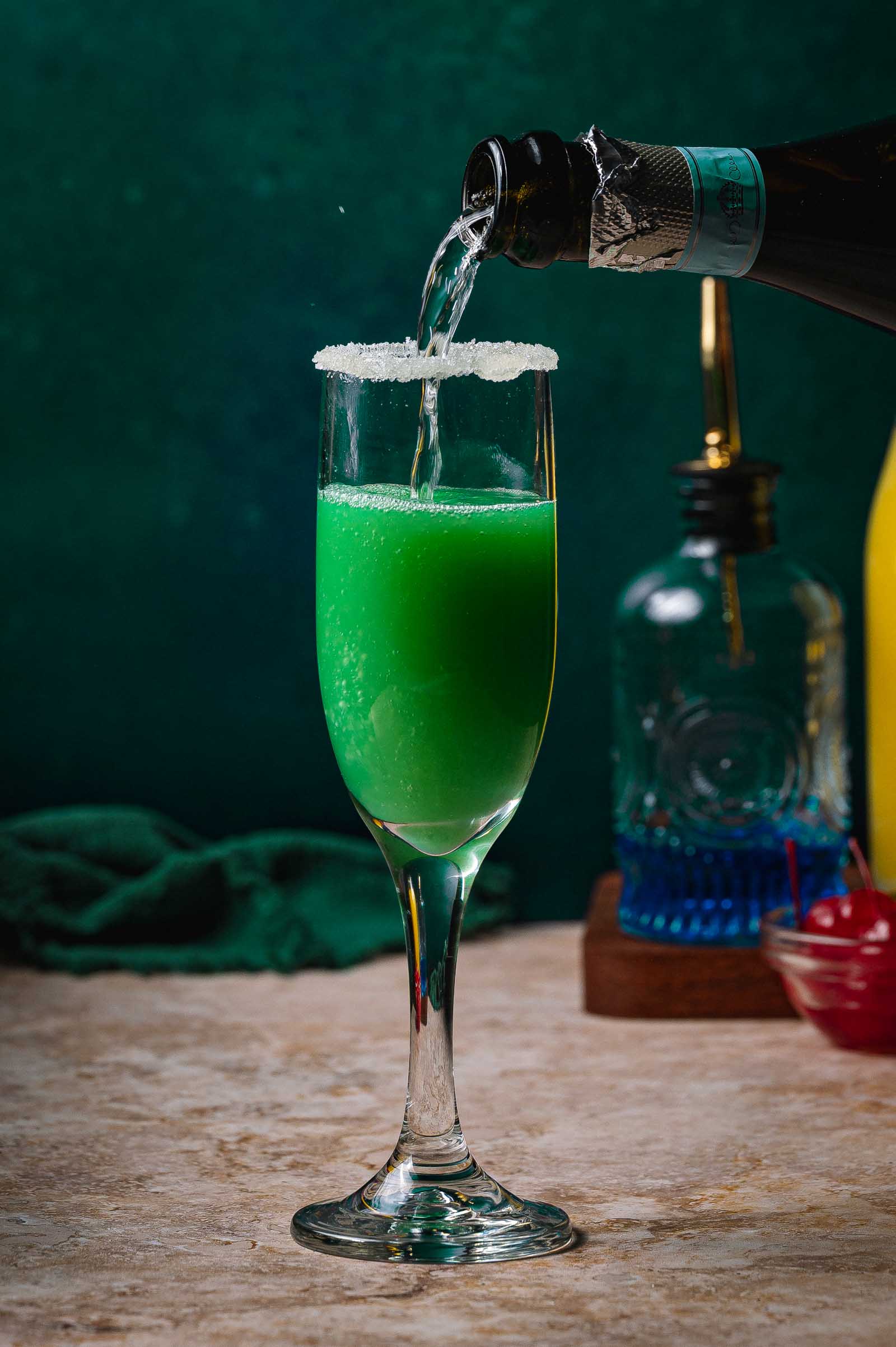 A sugar-rimmed flute holds a sparkling green Grinch mimosa cocktail as clear fizz is poured on top.