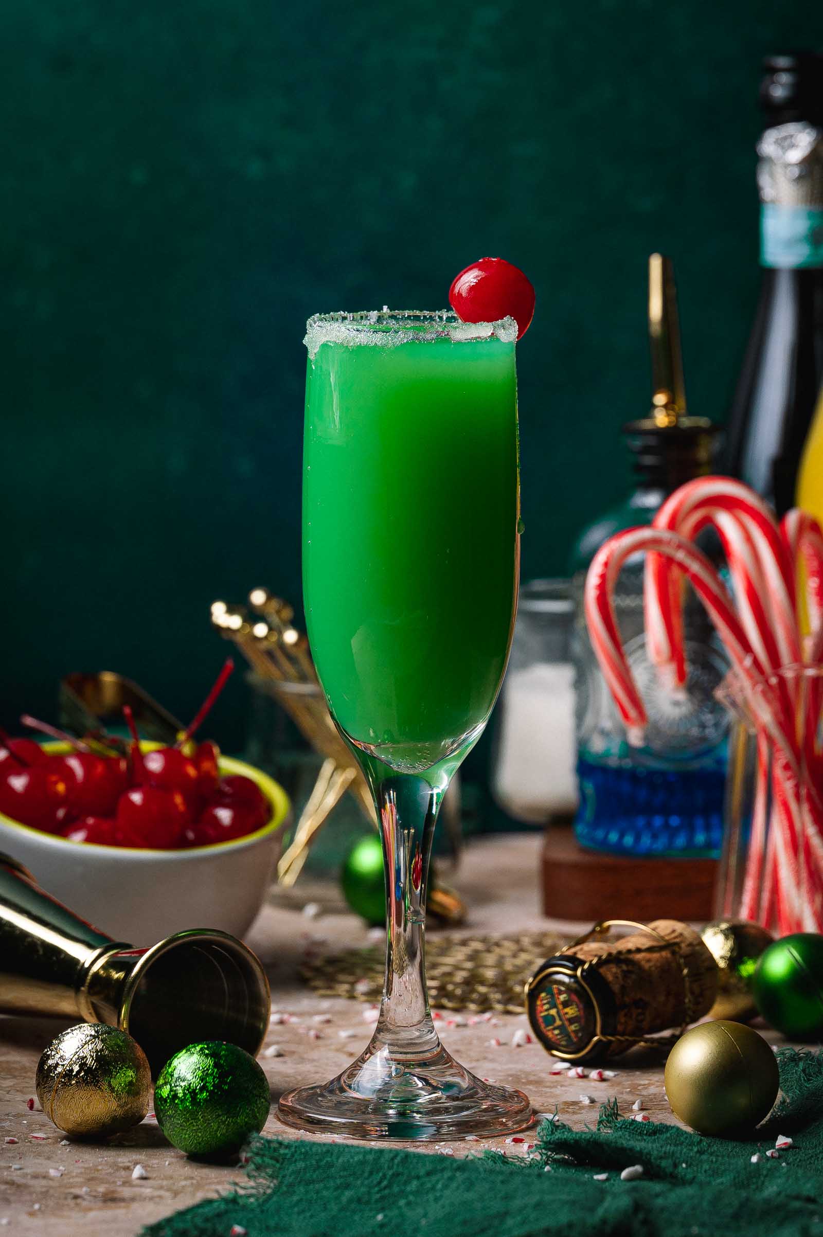 A green Grinch mimosa cocktail in a champagne flute with a red cherry, amid candy canes, cherries, gold ornaments, and a bottle behind.