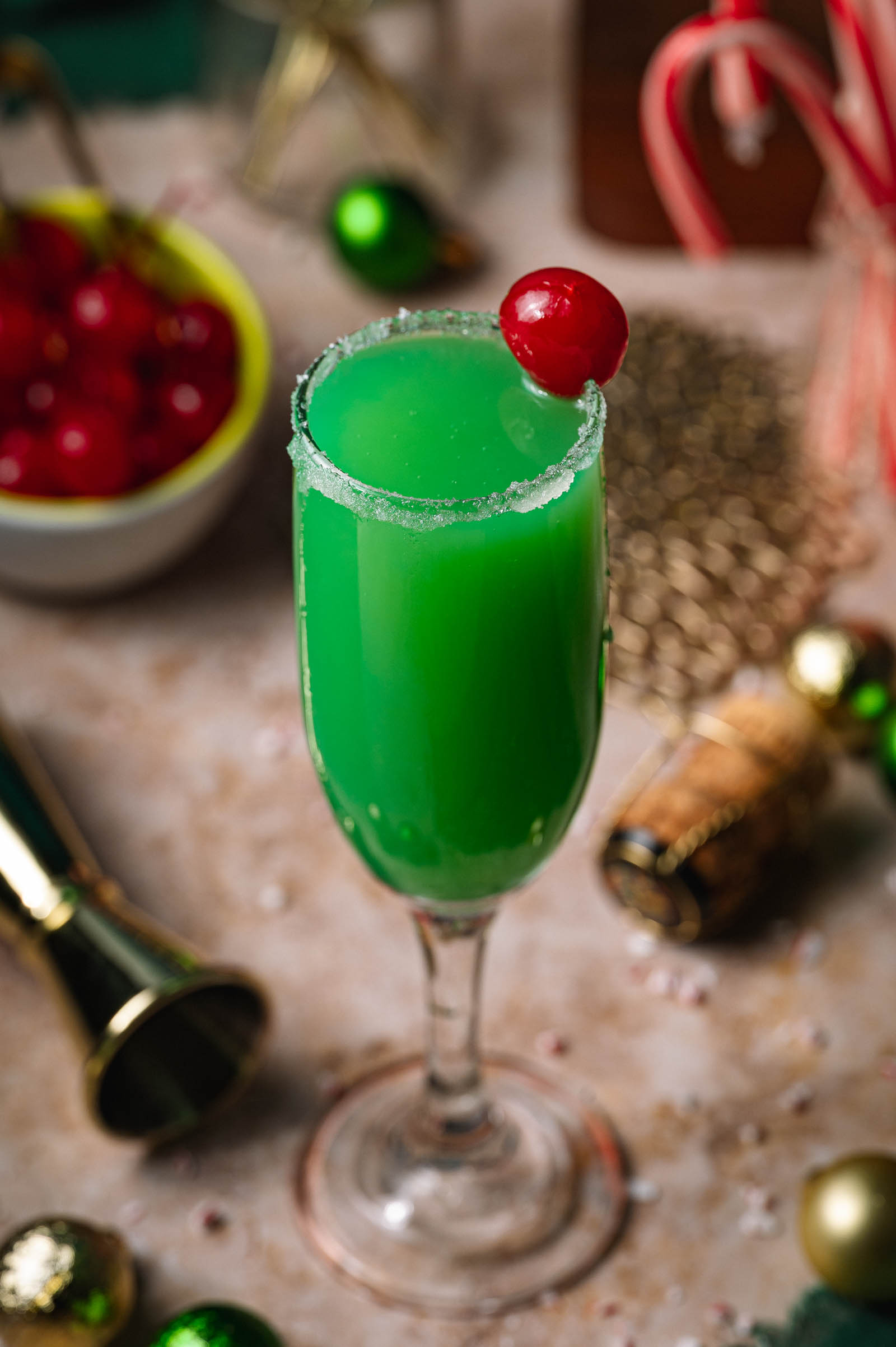 A green Grinch mimosa cocktail in a sugar-rimmed flute with a cherry garnish sits amid holiday decorations and drink ingredients.