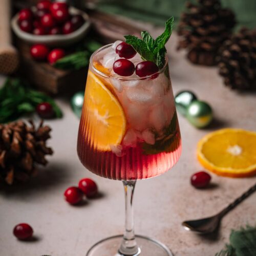 A wine glass with a Holiday Hugo Spritz, cranberries, orange slice, and mint sits among pinecones and holiday decor on a rustic table.