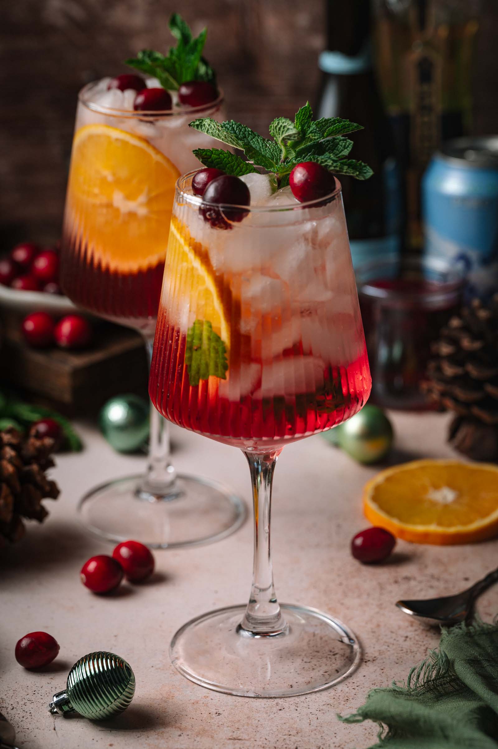 Two stemmed glasses of iced Holiday Hugo Spritz cocktails with orange slices, cranberries, and mint, surrounded by festive holiday decorations.