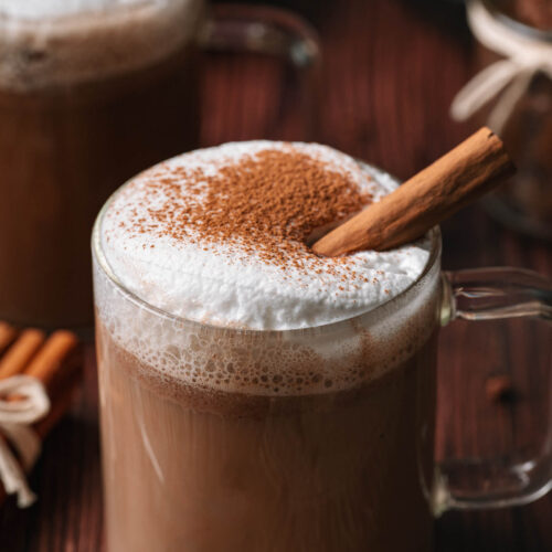 A glass mug of frothy hot buttered rum with cinnamon powder and a stick, alongside another similar mug on a wooden surface.