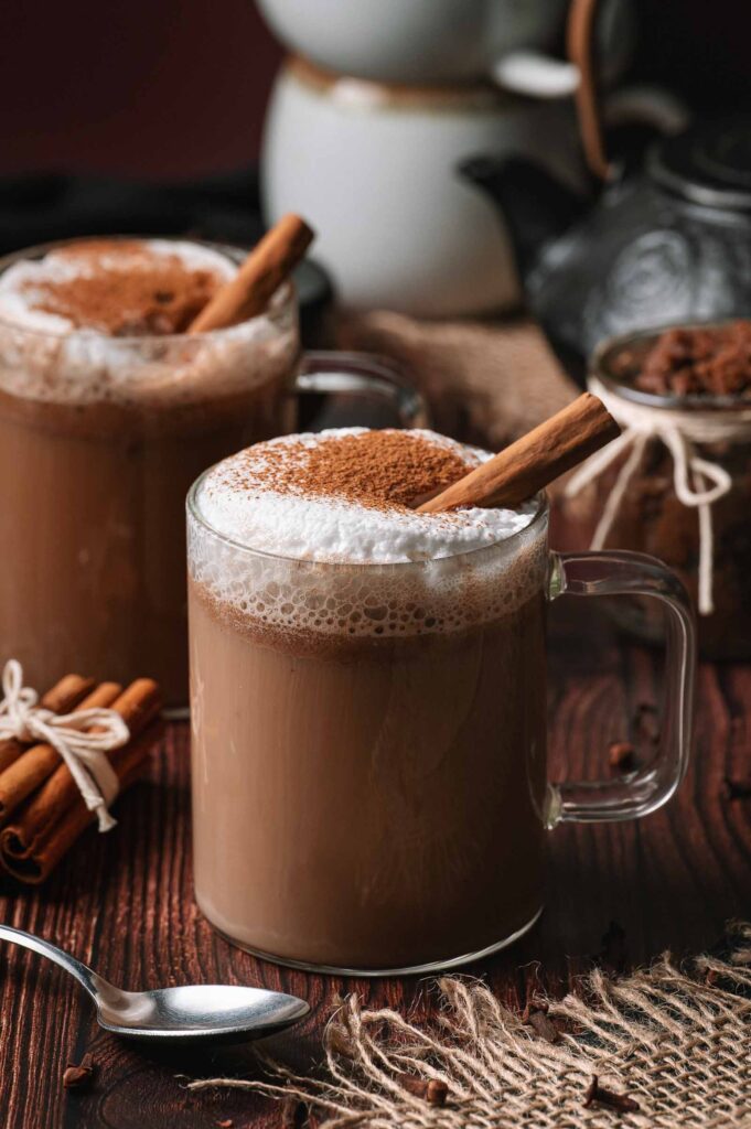 Two glass mugs of frothy hot buttered rum with cinnamon sticks sit on a wooden table, surrounded by coffee beans and a spoon.