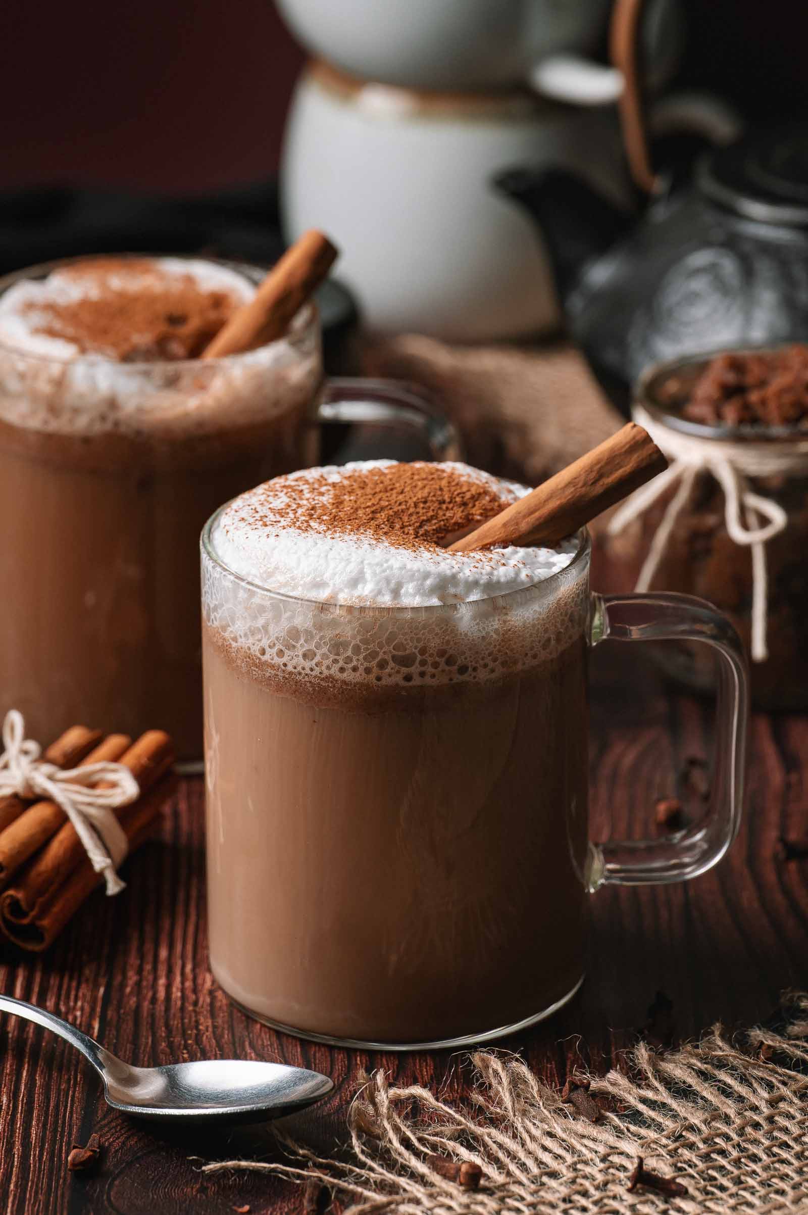 Two glass mugs of frothy hot buttered rum with cinnamon sticks sit on a wooden table, surrounded by coffee beans and a spoon.