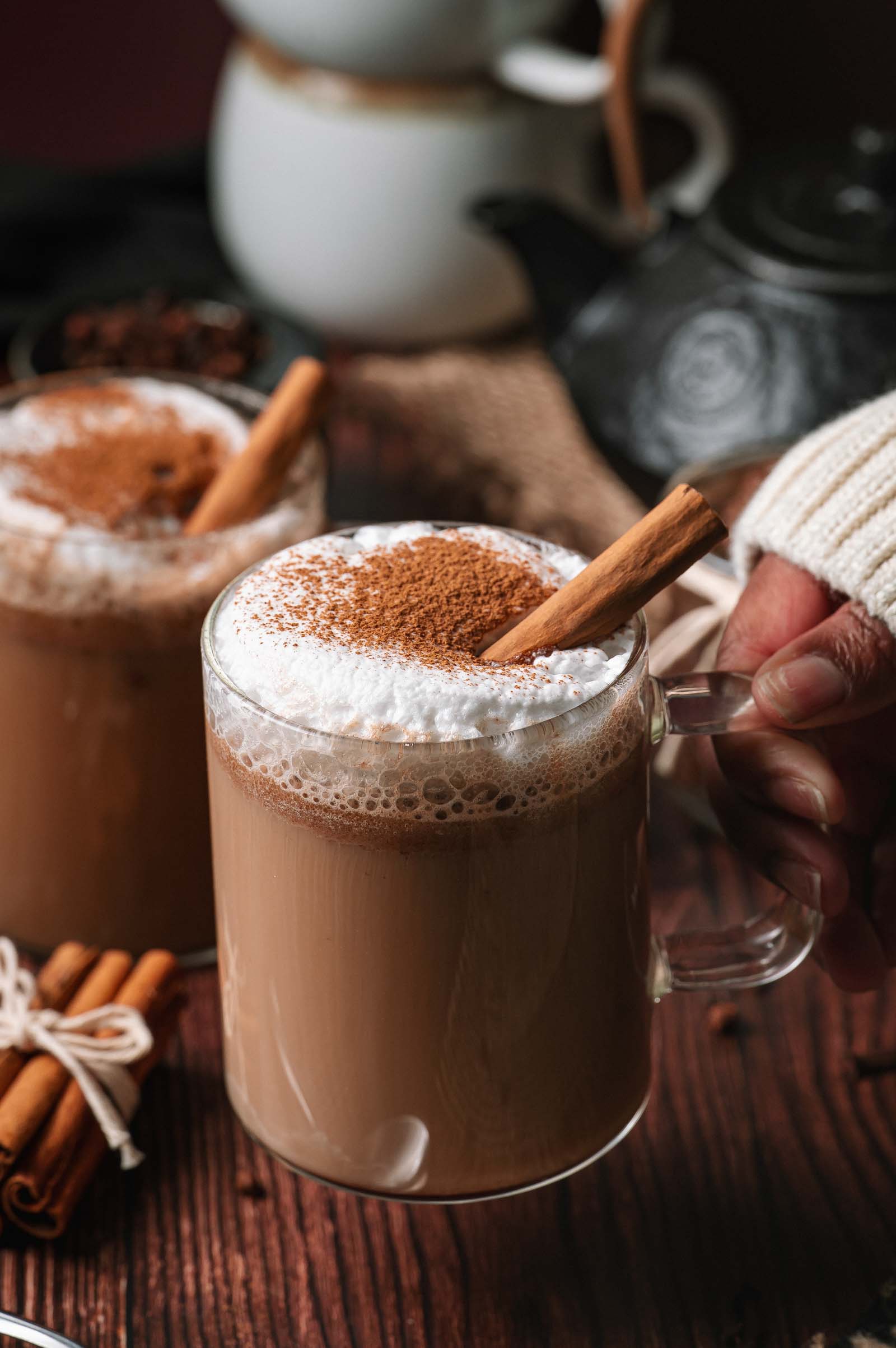 A hand holds a glass mug of frothy hot buttered rum topped with cinnamon and a stick, with another similar mug in the background.