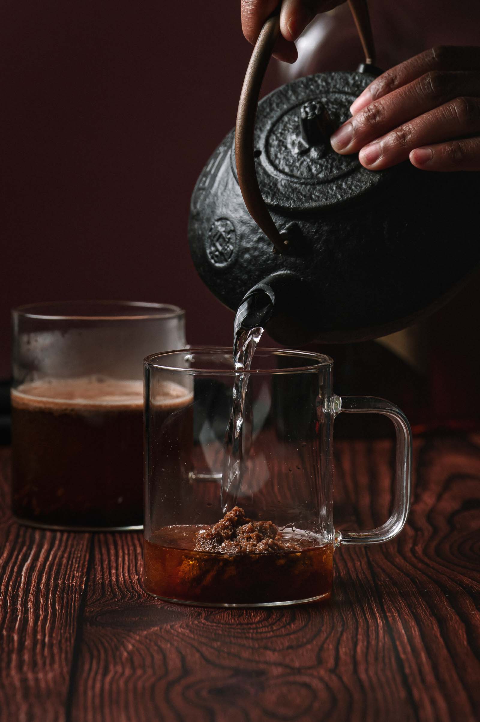 A person pours hot water from a black teapot into a glass mug of rum and hot buttered rum batter next to another filled glass on a wooden surface.