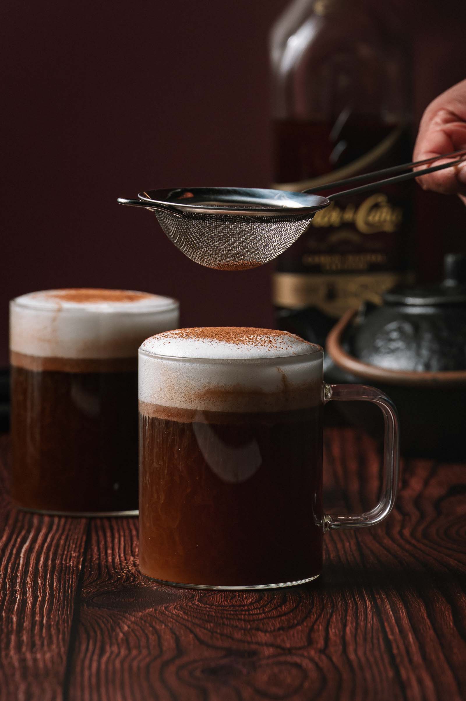 A person sifts cocoa powder over a glass of frothy hot buttered rum on a wooden table, with another mug and bottle in the background.