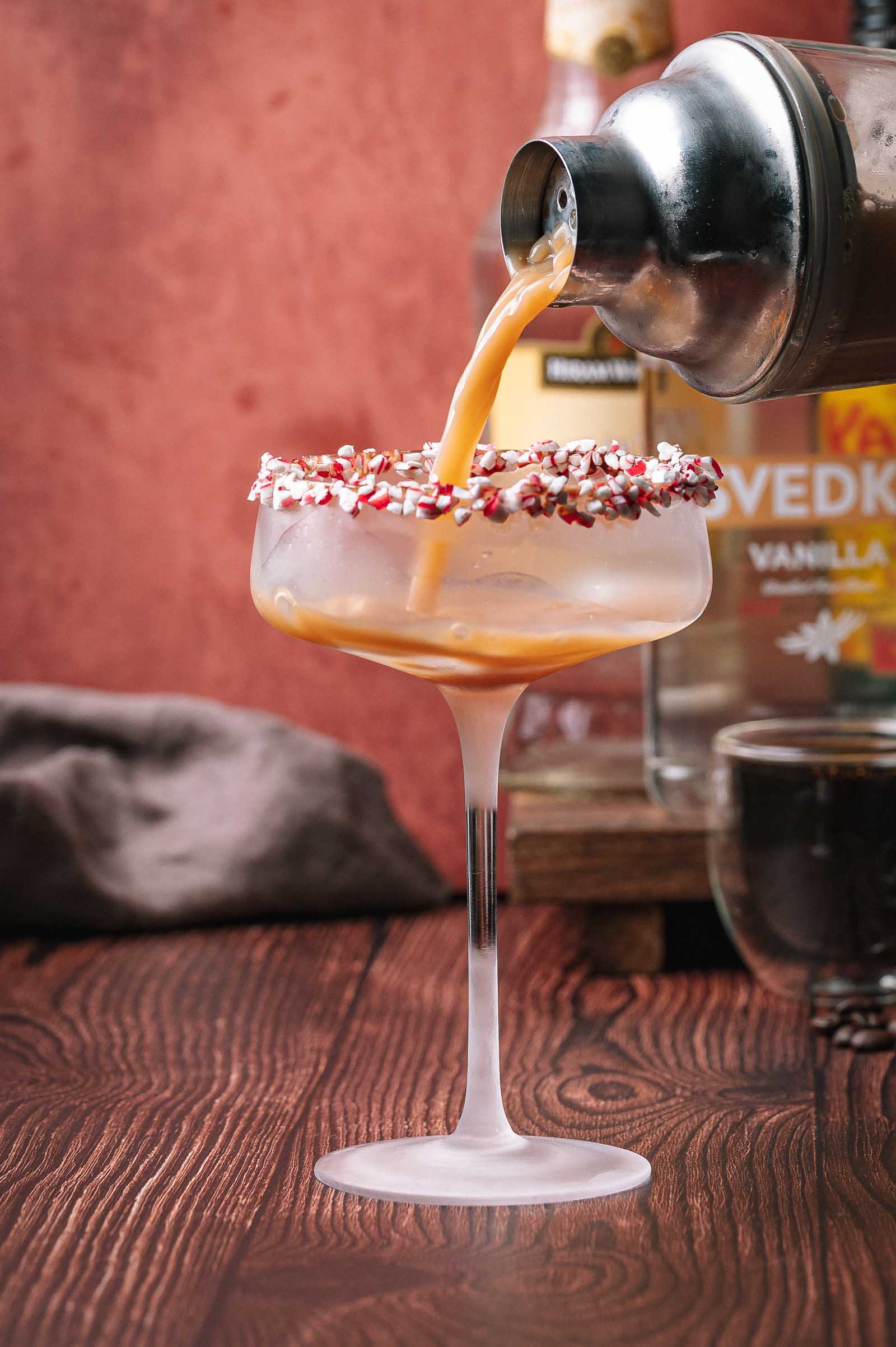 A peppermint mocha espresso martini cocktail pours from a shaker into a sprinkle-rimmed glass, with a “Vanilla Vodka” bottle in the background.
