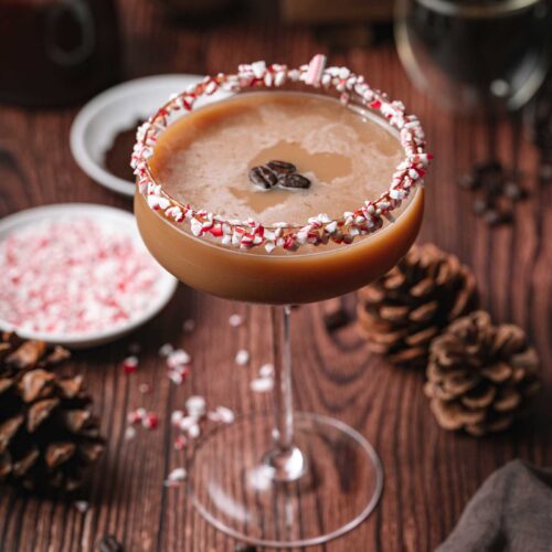 A coupe glass peppermint mocha espresso martini with a crushed peppermint rim and coffee beans sits on a wooden table with pinecones and crushed peppermint.