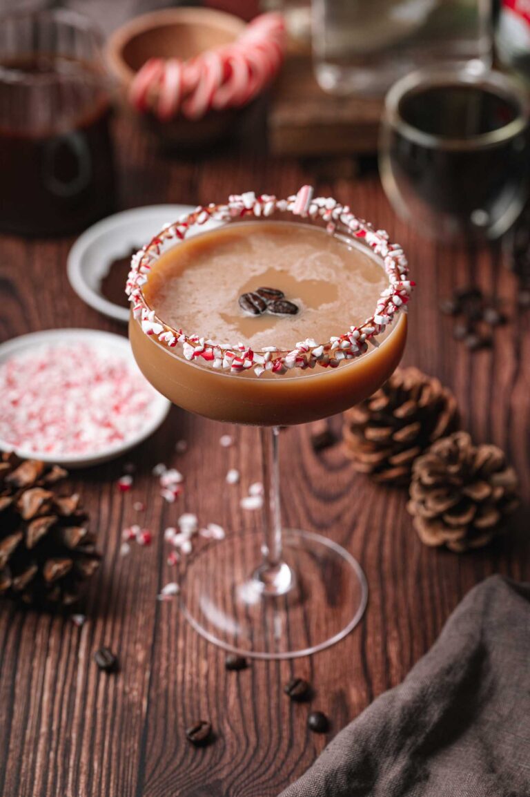 A coupe glass peppermint mocha espresso martini with a crushed peppermint rim and coffee beans sits on a wooden table with pinecones and crushed peppermint.