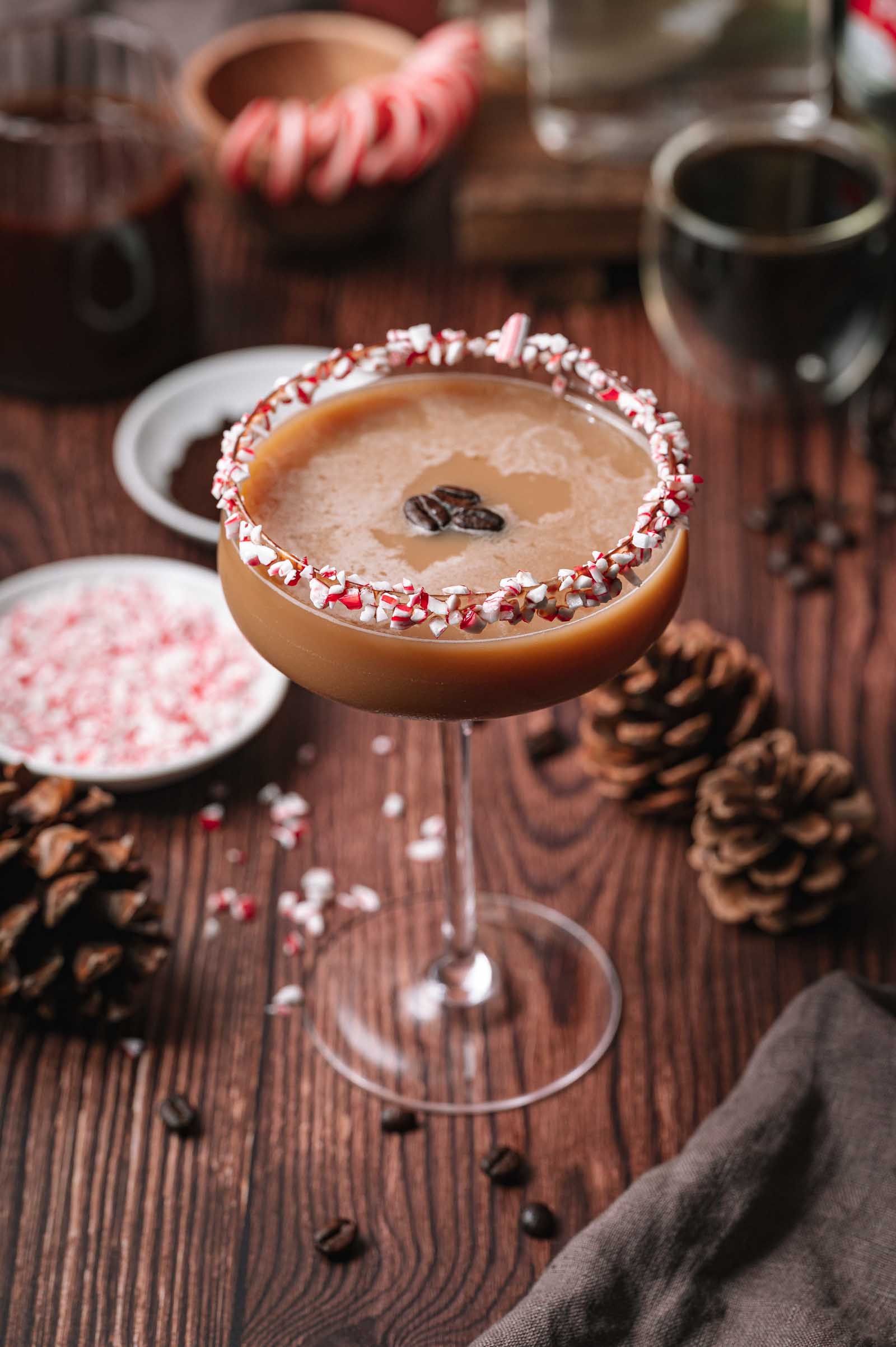 A coupe glass peppermint mocha espresso martini with a crushed peppermint rim and coffee beans sits on a wooden table with pinecones and crushed peppermint.
