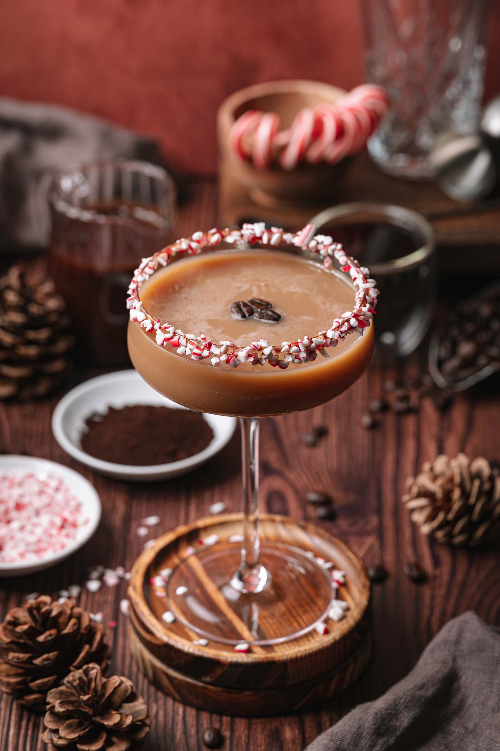Creamy peppermint mocha espresso martini in a coupe glass with a crushed candy cane rim and coffee beans, on a table with pinecones and holiday decor.