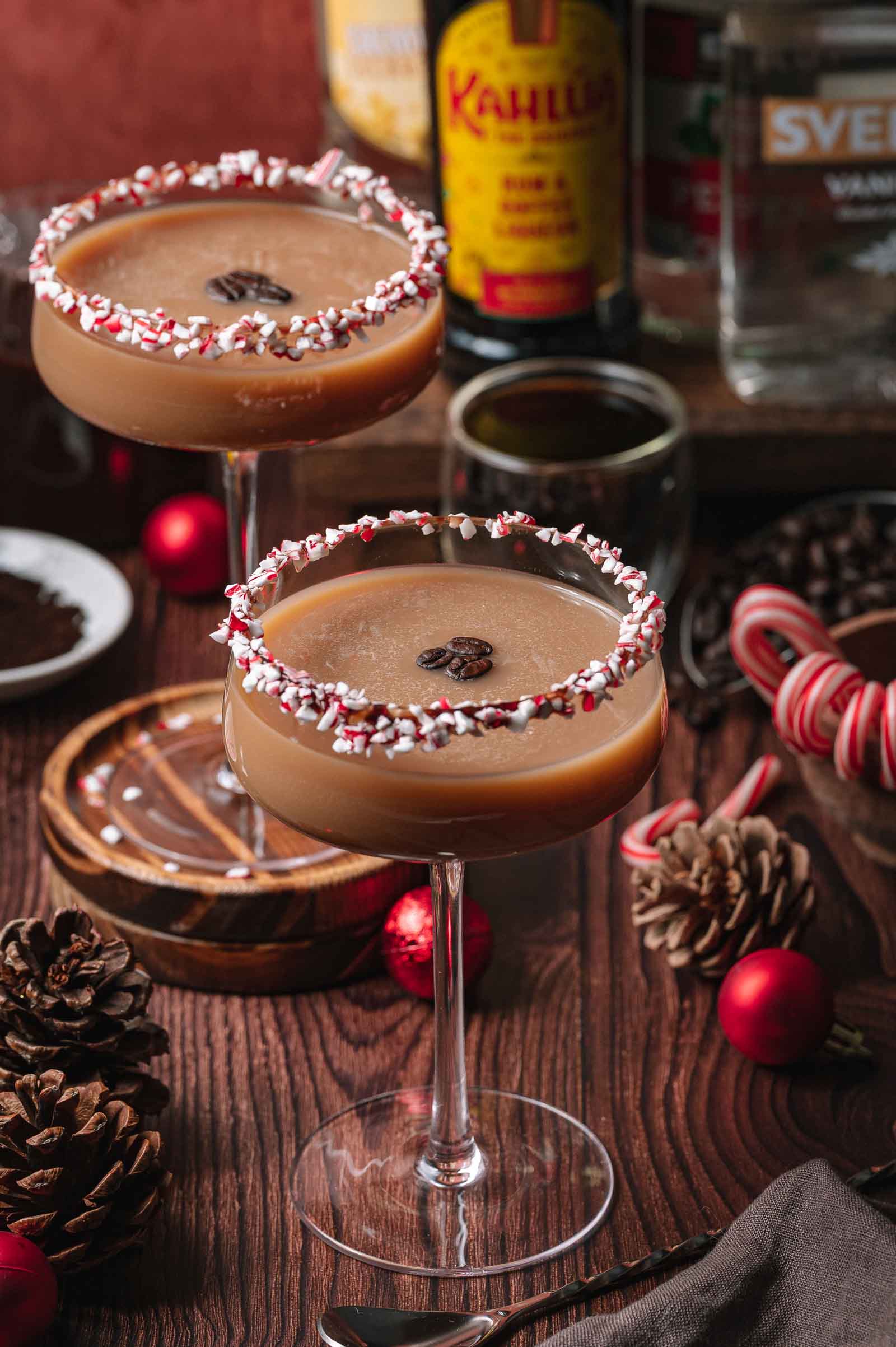 Two espresso martinis with peppermint rims sit on a wooden table amid pinecones, candy canes, ornaments, and liqueur bottles.