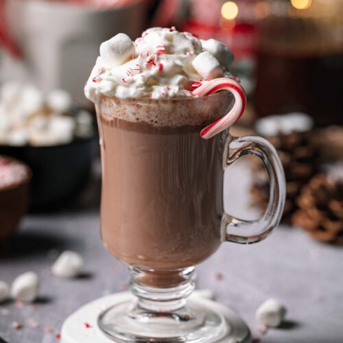 Spiked Peppermint Hot Chocolate in a glass mug with whipped cream, marshmallows, crushed peppermint, and a candy cane on a table.
