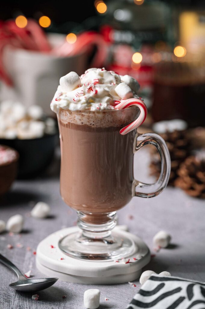 Spiked Peppermint Hot Chocolate in a glass mug with whipped cream, marshmallows, crushed peppermint, and a candy cane on a table.