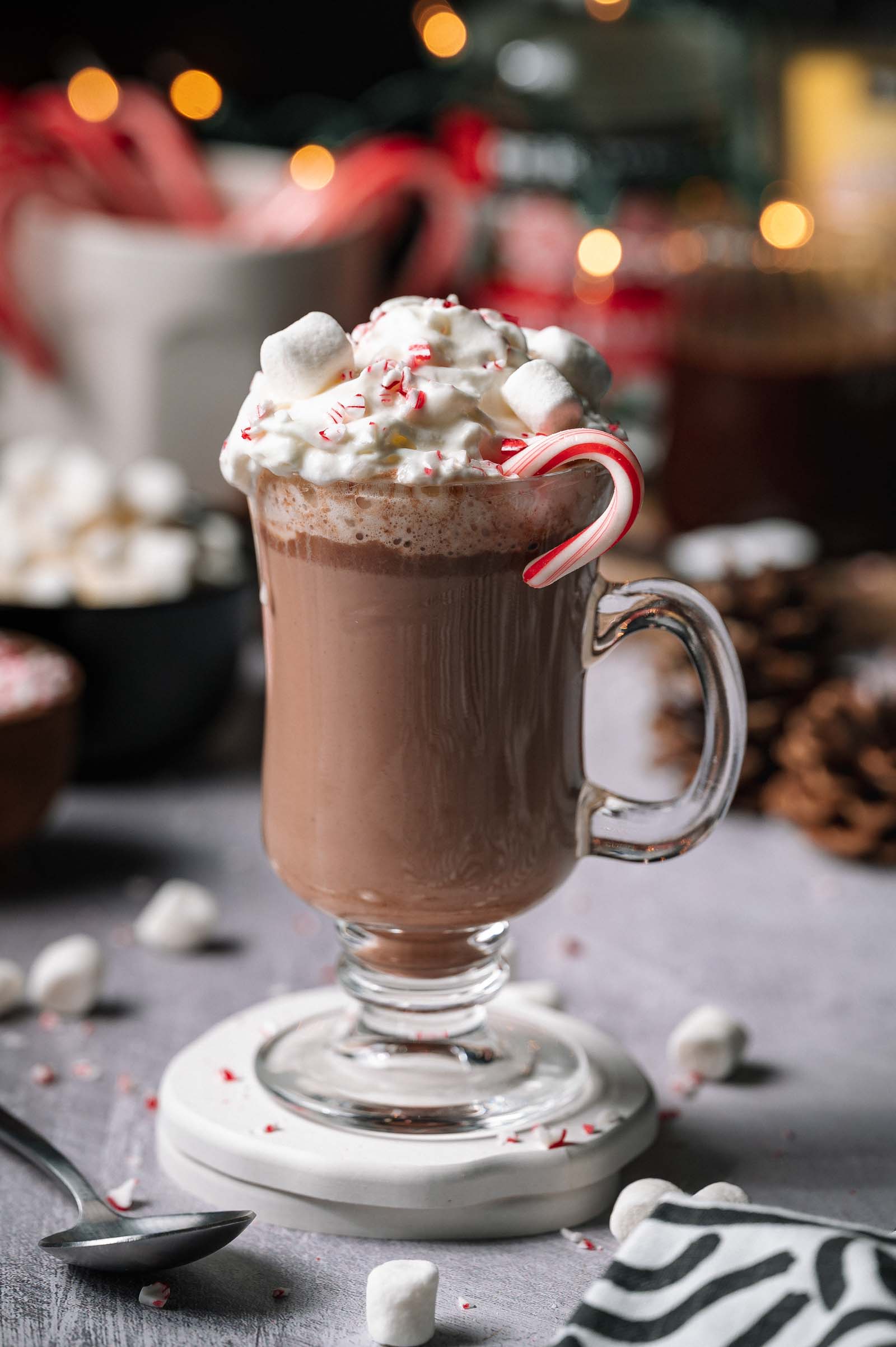 Spiked Peppermint Hot Chocolate in a glass mug with whipped cream, marshmallows, crushed peppermint, and a candy cane on a table.