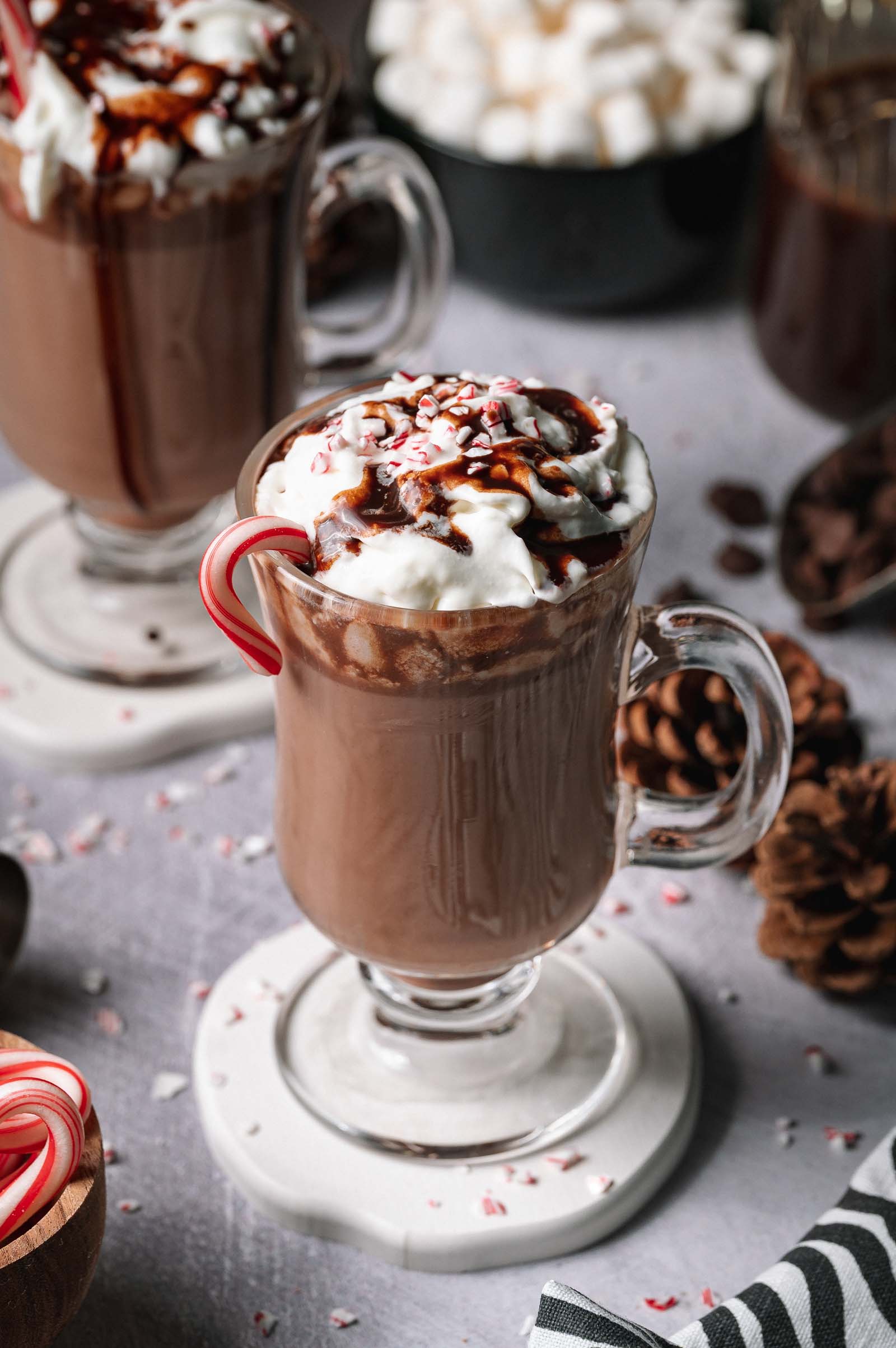 A glass mug of Spiked Peppermint Hot Chocolate with whipped cream, chocolate syrup, peppermint, and a candy cane, plus another mug behind it.