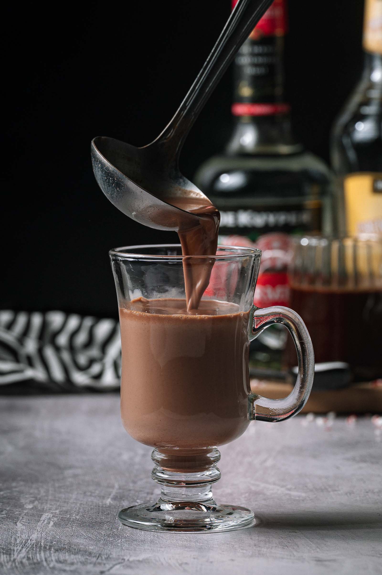 A ladle pours spiked hot chocolate into a clear glass mug, with liquor bottles and a patterned cloth in the background.