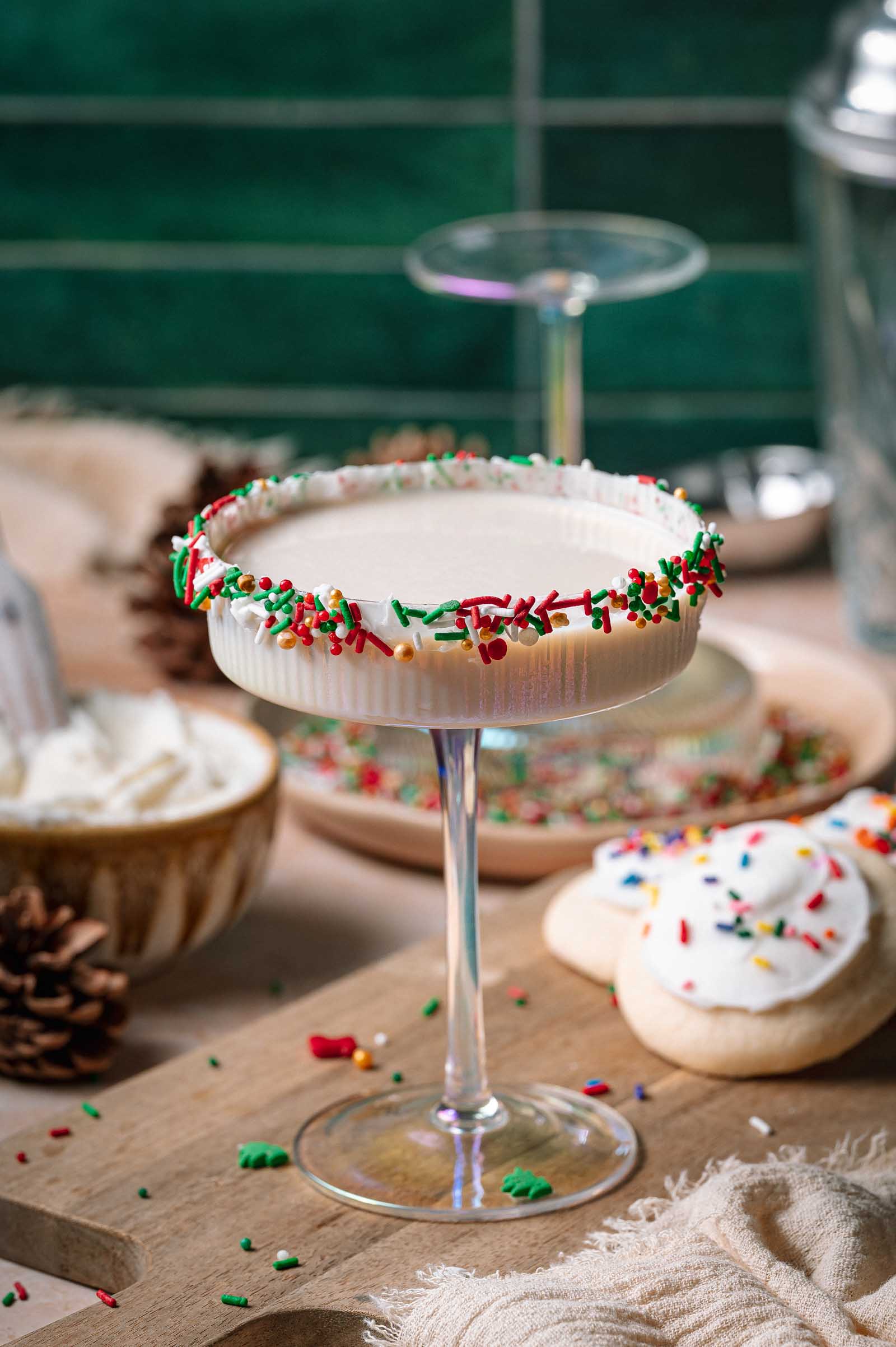 A sprinkle-rimmed sugar cookie martini cocktail sits on a wooden board alongside frosted sugar cookies and a bowl of whipped cream.