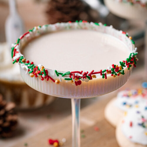 A creamy sugar cookie martini in a sprinkle-rimmed coupe glass sits with pinecones and a frosted cookie in the background.