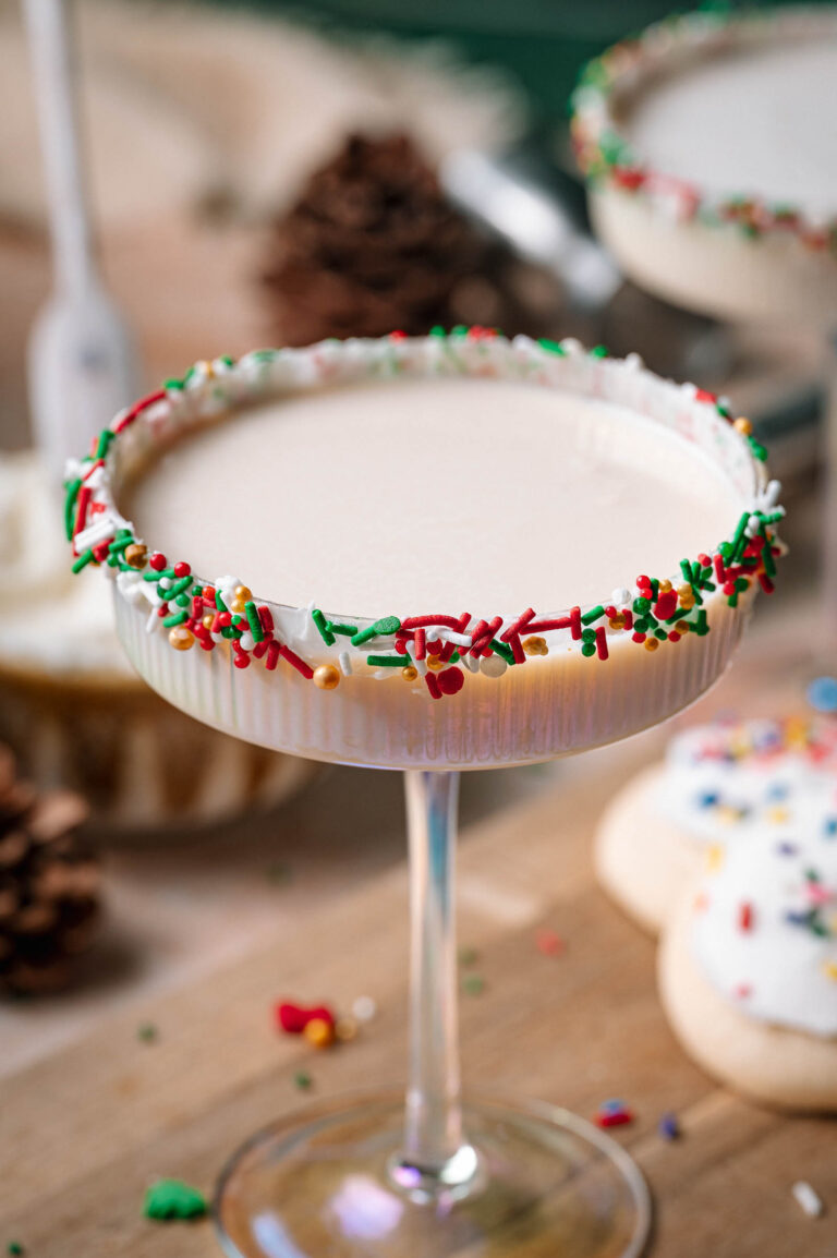 A creamy sugar cookie martini in a sprinkle-rimmed coupe glass sits with pinecones and a frosted cookie in the background.