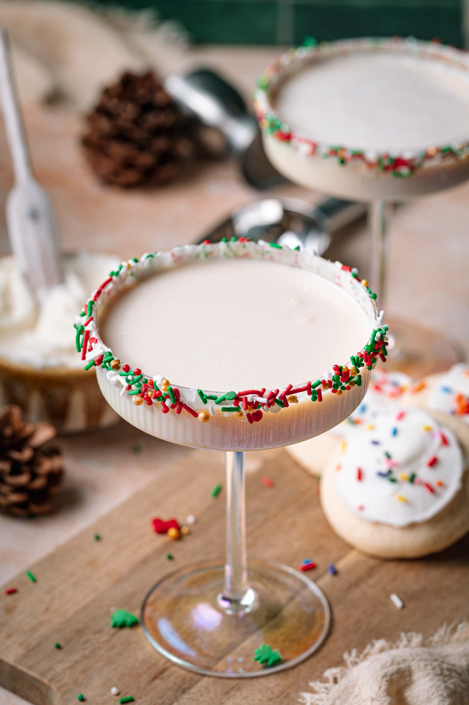 A creamy sugar cookie martini cocktail in a sprinkle-rimmed glass sits on a wooden board with frosted cookies and pinecones in the background.