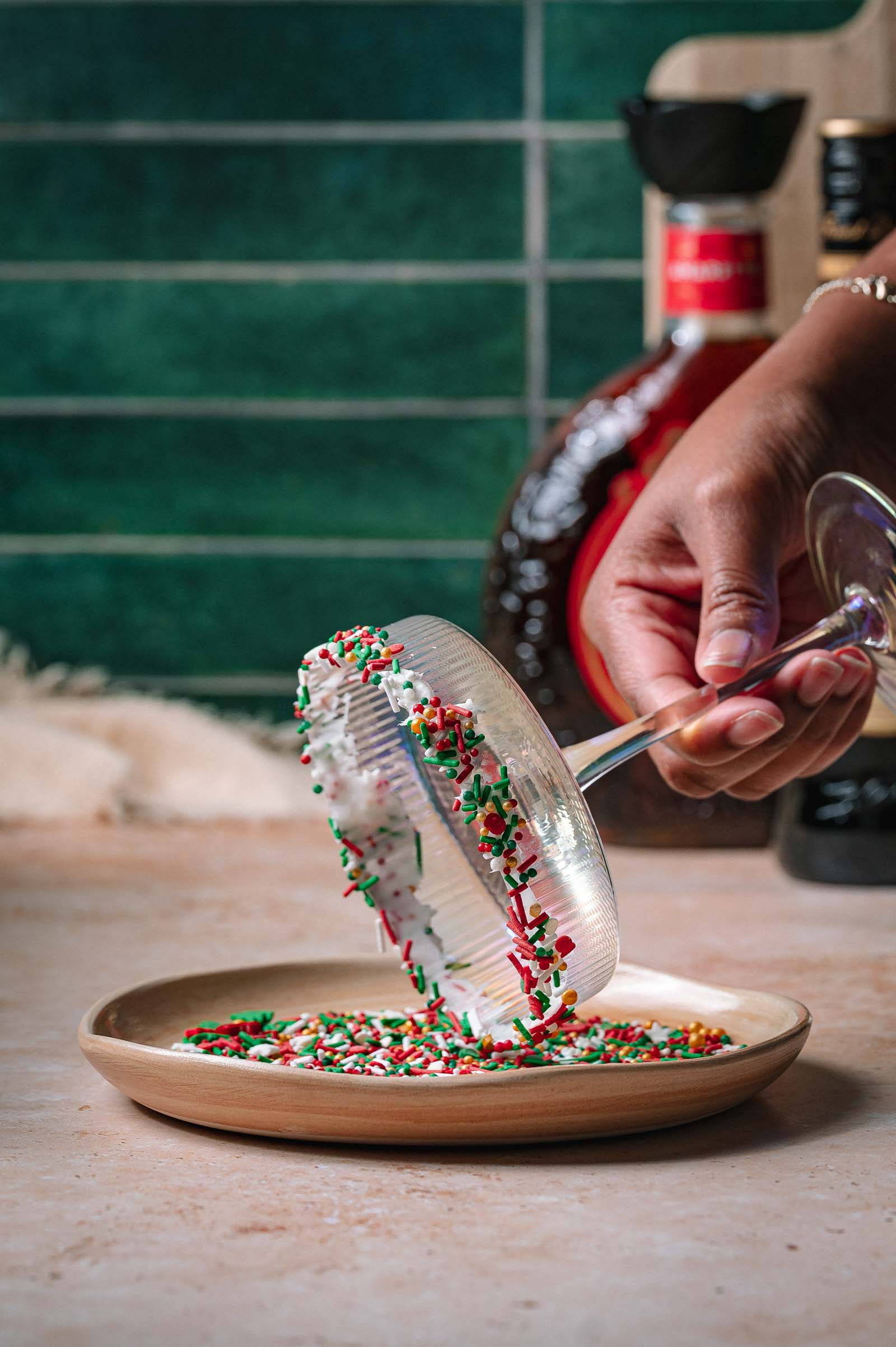 A hand dips a cocktail glass rim into red, green, and white sprinkles, with bottles and a green tiled wall behind.