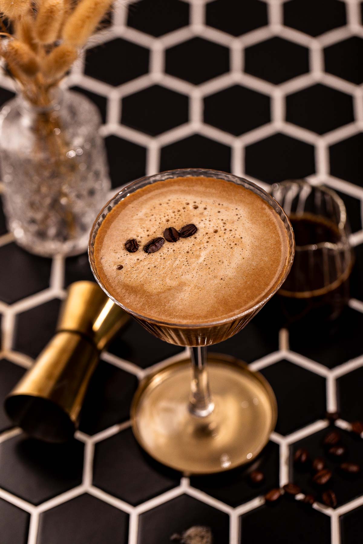 An espresso martini in a stemmed glass, garnished with three coffee beans, sits on a black-and-white hexagonal tile surface.