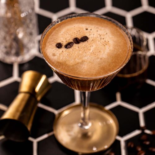 An espresso martini in a ribbed coupe glass garnished with three coffee beans sits on a black hexagonal tiled surface next to a gold jigger.