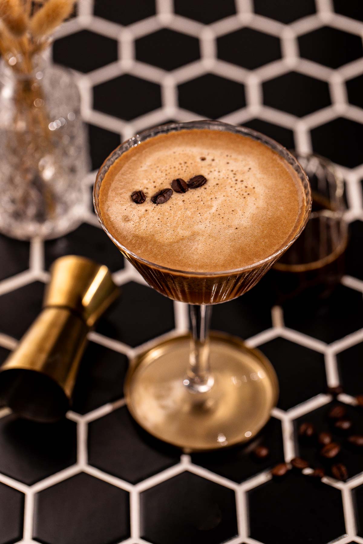 An espresso martini in a ribbed coupe glass garnished with three coffee beans sits on a black hexagonal tiled surface next to a gold jigger.