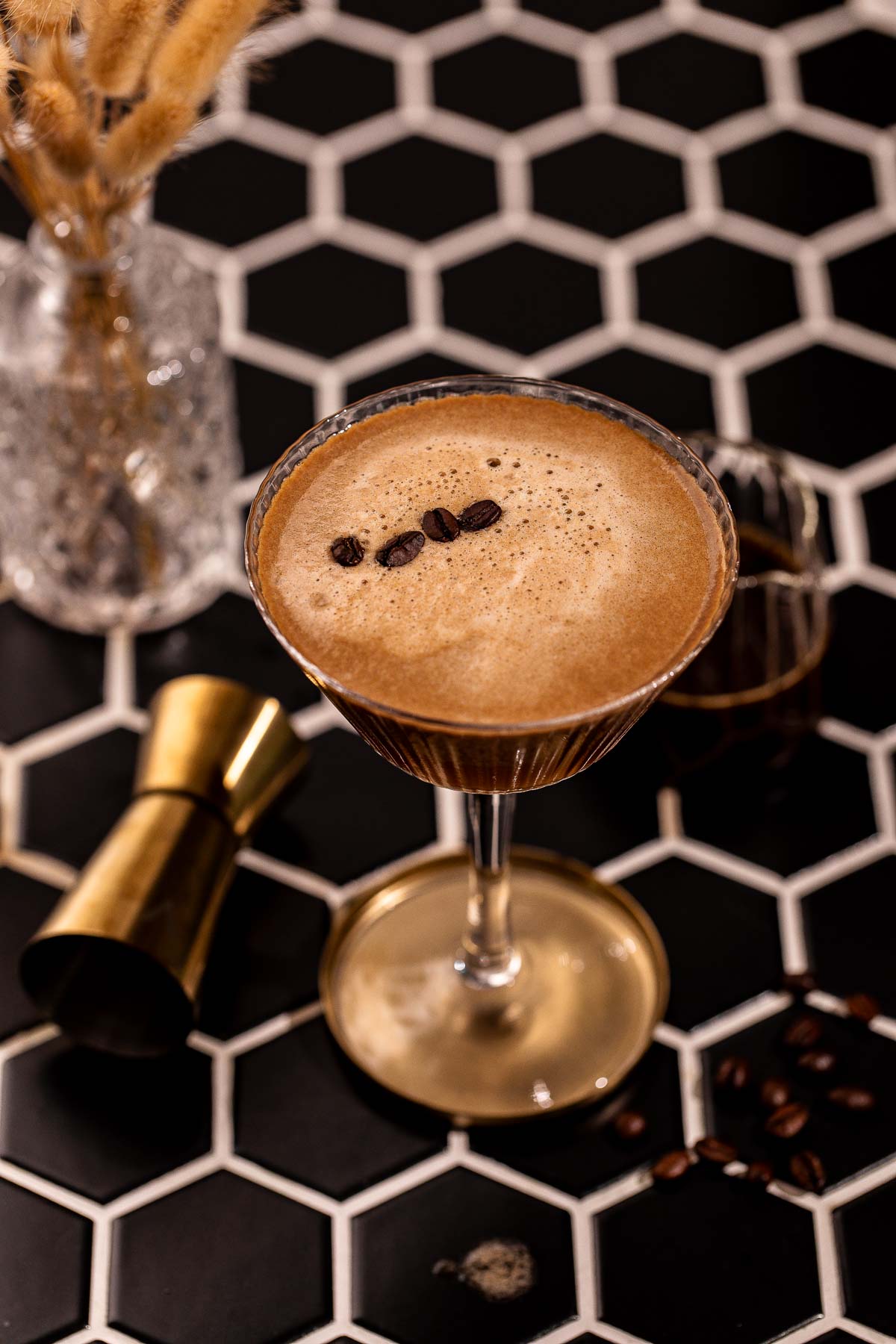 An espresso martini in a stemmed glass garnished with three coffee beans sits on a black and white hexagonal tile surface.