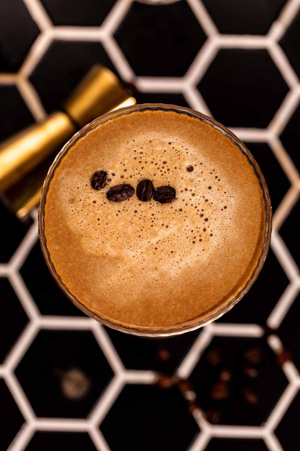 An espresso martini topped with three coffee beans rests on a hexagonal tile surface.