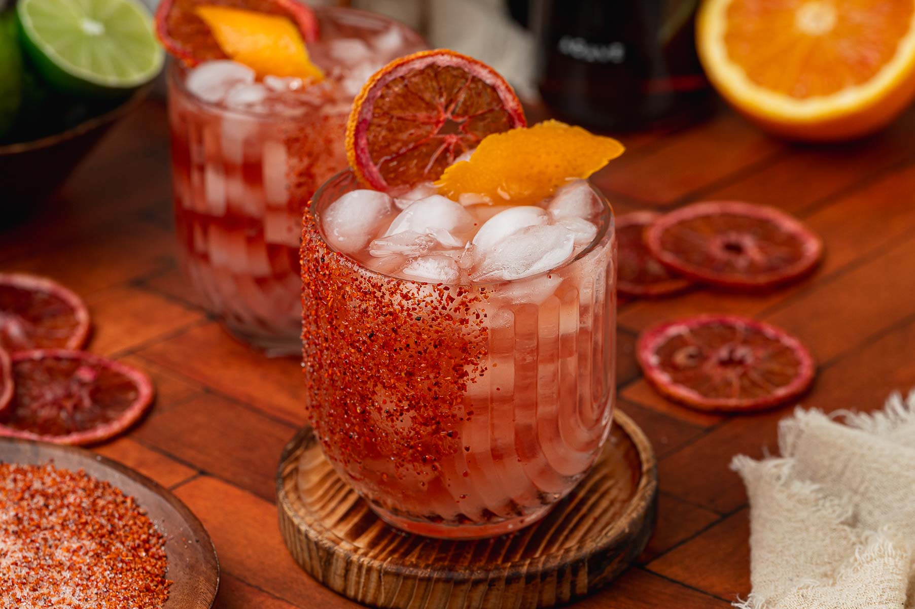 Two chili-salt rimmed glasses of reddish cocktail with ice, garnished with dried blood orange slices and citrus peel on a wooden table.