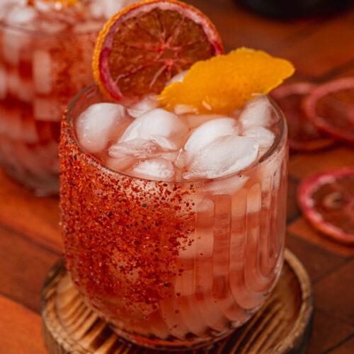 Two iced pink cocktails with chili-salted rims, garnished with dried blood orange and peel on wooden coasters.