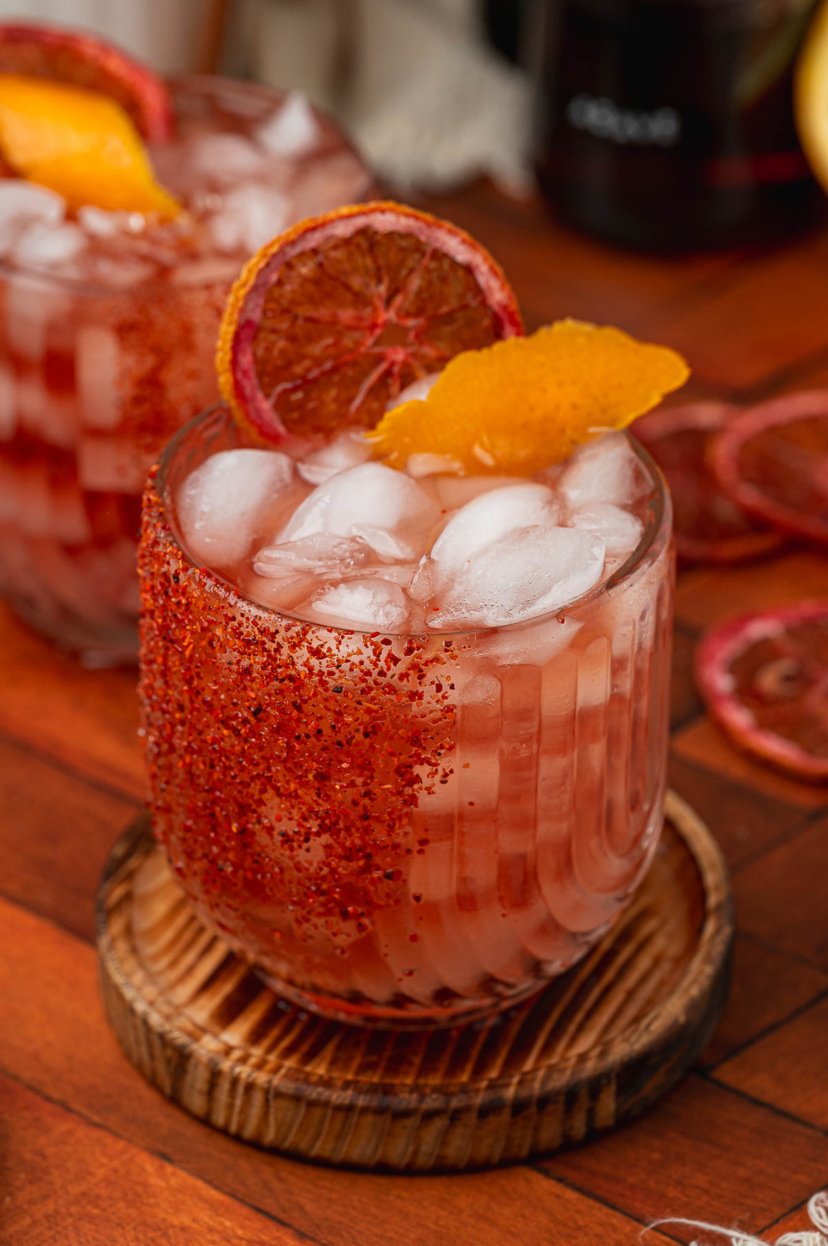 Two iced pink cocktails with chili-salted rims, garnished with dried blood orange and peel on wooden coasters.