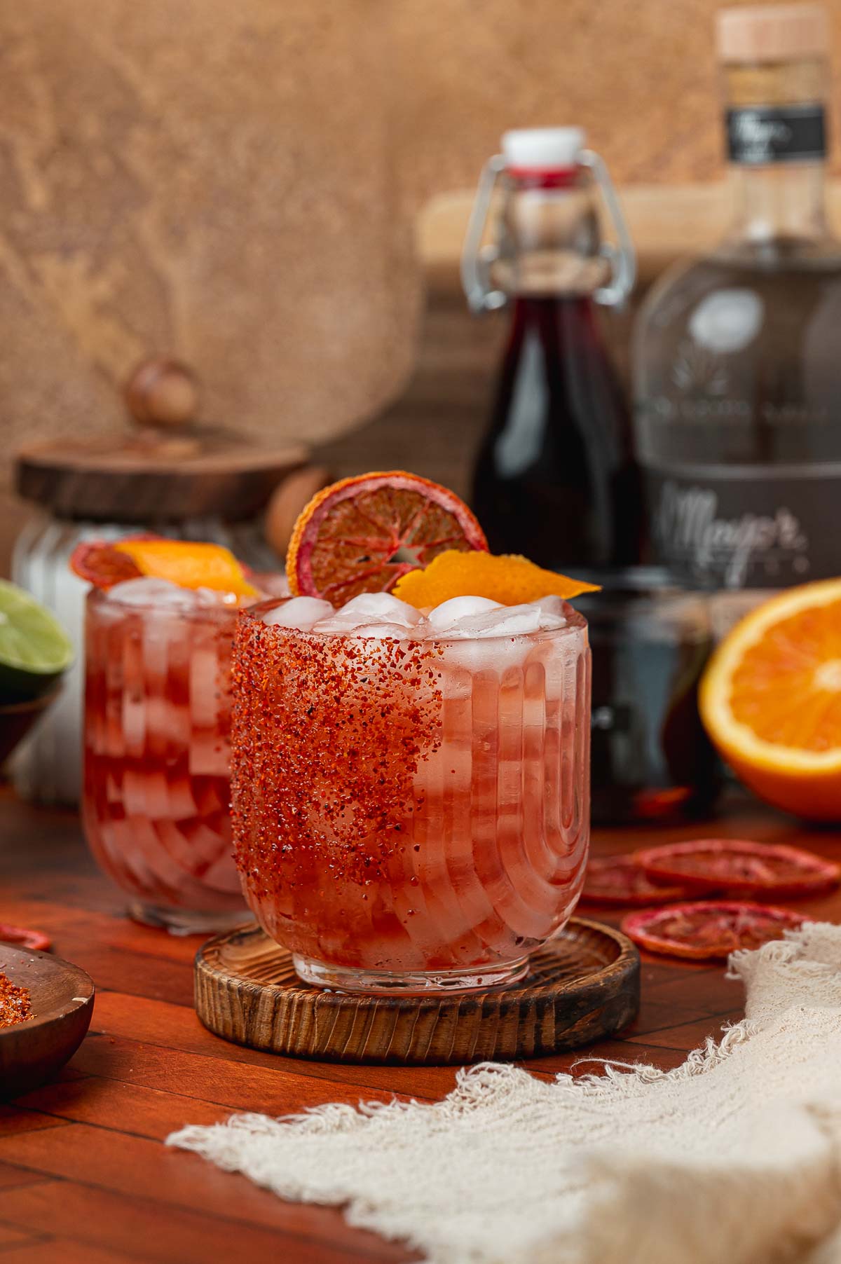 Two reddish cocktails with ice, chili-salt rims, and dehydrated orange slice garnishes.